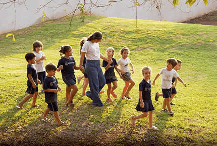 Grupo de crianças caminhando ao ar livre em um gramado, acompanhadas por uma professora, durante uma atividade escolar.