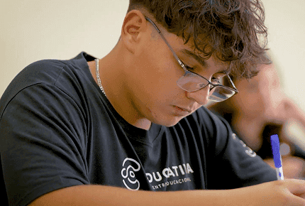 Estudante concentrado escrevendo em sala de aula, usando camiseta da Educativa e óculos, durante um simulado.